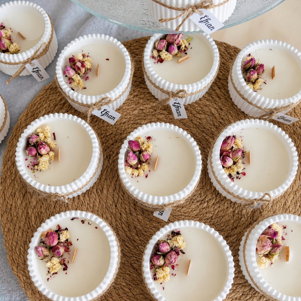 A styled flat-lay of multiple small white candles in ribbed white cups, arranged on a round woven mat. Each candle is topped with dried pink rose petals, yellow flower pieces, and a small wooden wick.