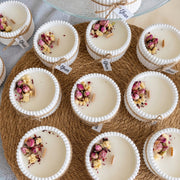 A styled flat-lay of multiple small white candles in ribbed white cups, arranged on a round woven mat. Each candle is topped with dried pink rose petals, yellow flower pieces, and a small wooden wick.