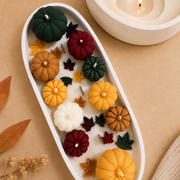 A styled flat-lay of small decorative pumpkins and autumn leaves arranged in a white oval tray on a tan surface, with a lit candle and dried leaves nearby.
