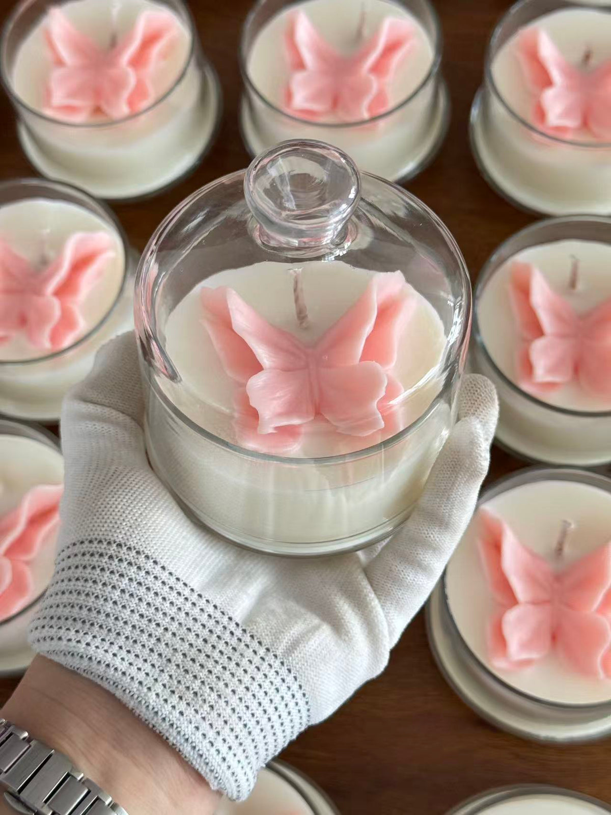 A hand wearing a white knit glove holds a clear glass jar candle with white wax and a pink butterfly ornament inside. Multiple similar butterfly candles are arranged on a wooden surface in the background.