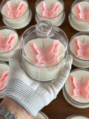 A hand wearing a white knit glove holds a clear glass jar candle with white wax and a pink butterfly ornament inside. Multiple similar butterfly candles are arranged on a wooden surface in the background.
