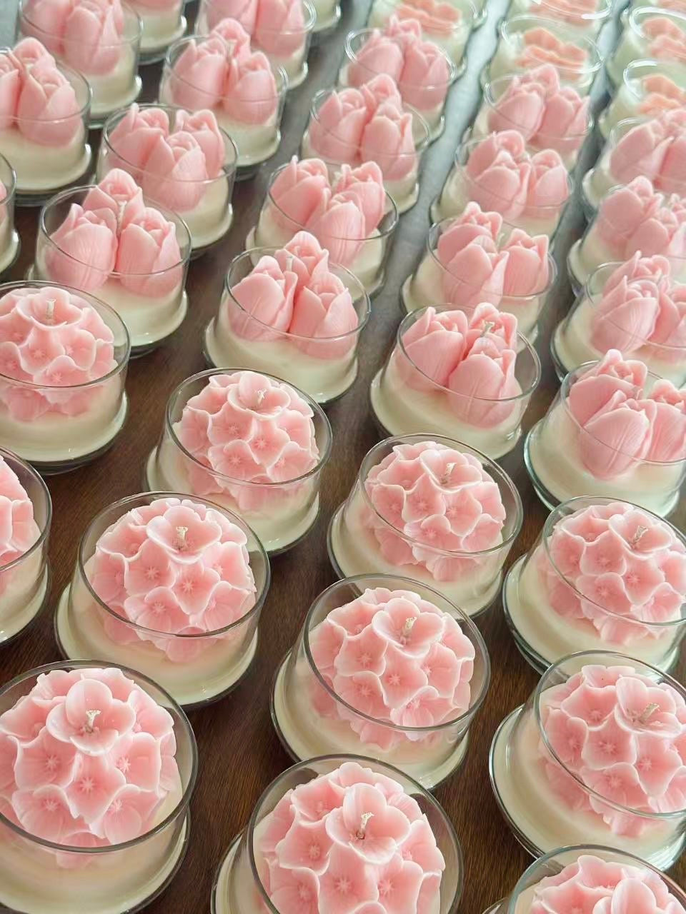 Multiple pink flower-shaped candles in clear glass holders arranged in rows on a wooden surface.
