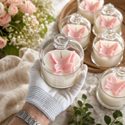 A hand wearing a white knit glove holds a small clear glass jar candle with a pink butterfly decoration on top. Several matching glass jar candles with butterfly decorations are arranged on a wooden tray and a light fabric surface, with pink flowers and green leaves in the background.