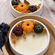 A round white candle in a light wood tray, decorated with three small decorative pumpkins in orange, dark green, and burgundy, along with orange and yellow floral accents. A matching second candle is visible in the background on a pale pink fabric surface.