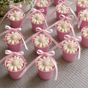 Multiple small pink bucket-shaped containers filled with cream flower-shaped wax melts or soaps, each tied with a pink ribbon, arranged on a textured beige fabric surface.