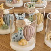 A set of clear glass candle holders containing layered white wax bases with whimsical animal and hot air balloon decorations, arranged on a wooden tabletop.