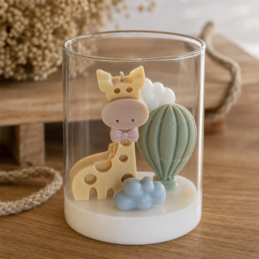 A clear cylindrical glass jar candle with a white wax base, decorated with a cute giraffe, hot air balloon, clouds, and a small blue cloud figure on a wooden surface.